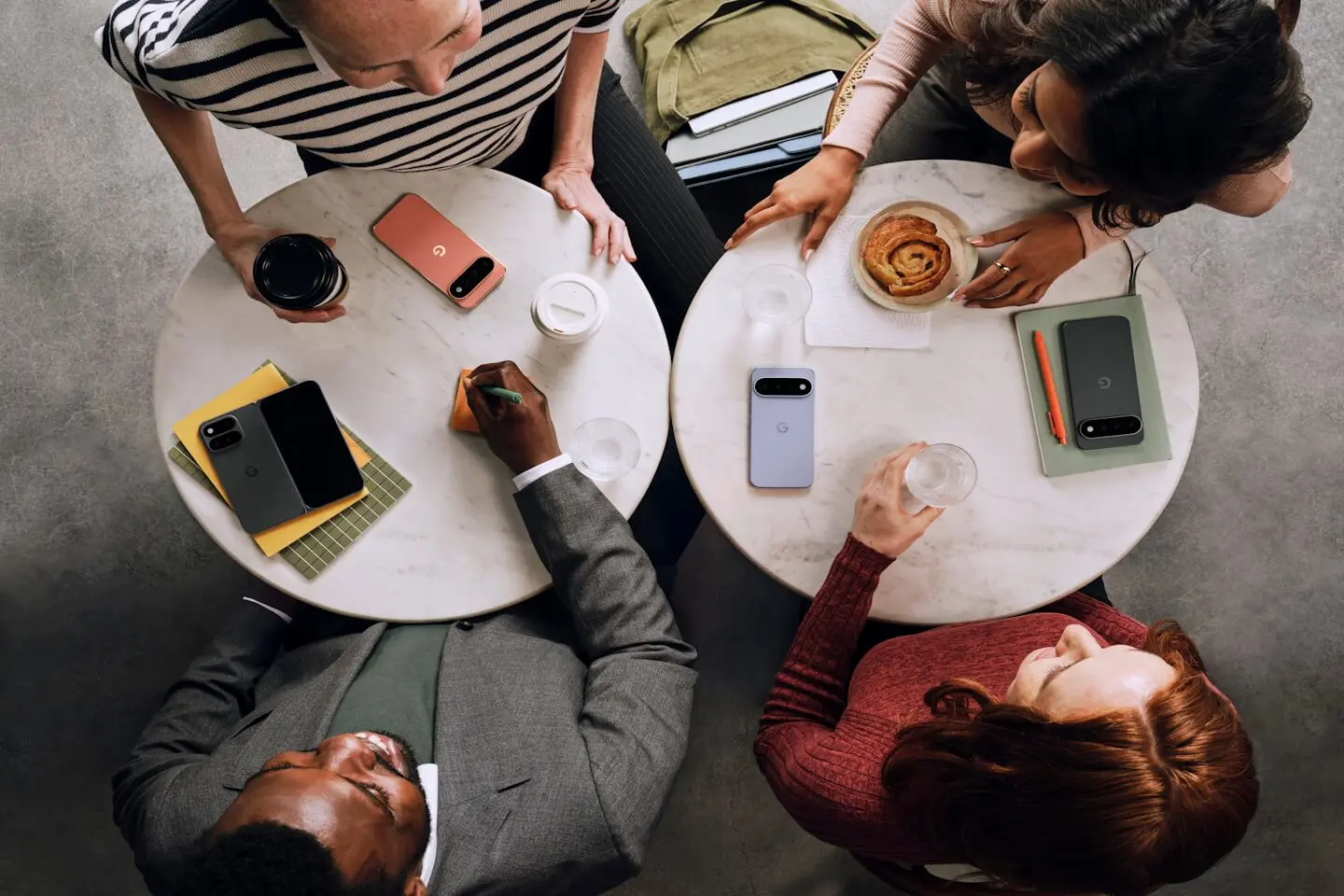 „Keturi žmonės sėdi kavinėje prie dviejų apvalių marmurinių stalų, ant kurių padėti skirtingų spalvų „Google Pixel 11“ telefonai, užrašų knygelės, gėrimai ir bandelė – vaizdas iš viršaus perteikia neformalų susitikimą ar pokalbį.