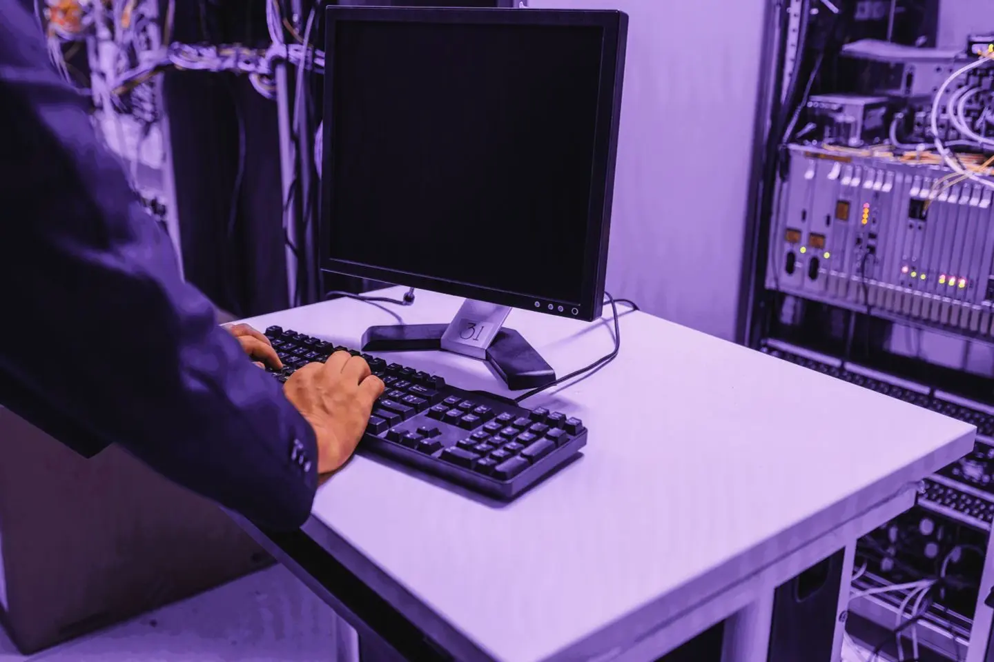 Asmuo dirba prie kompiuterio serverinėje, apšviestoje „Telia“ violetiniais atspalviais, tarp serverių ir tinklo įrangos.