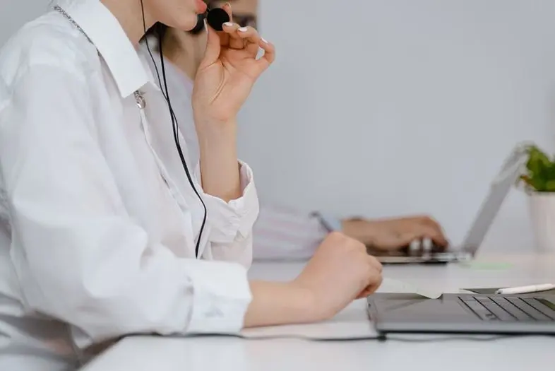 Biuro aplinkoje asmuo kalba per mikrofoną ir dirba kompiuteriu. Šalia yra užrašų priemonės, fone matomas kitas darbo vietos naudotojas.