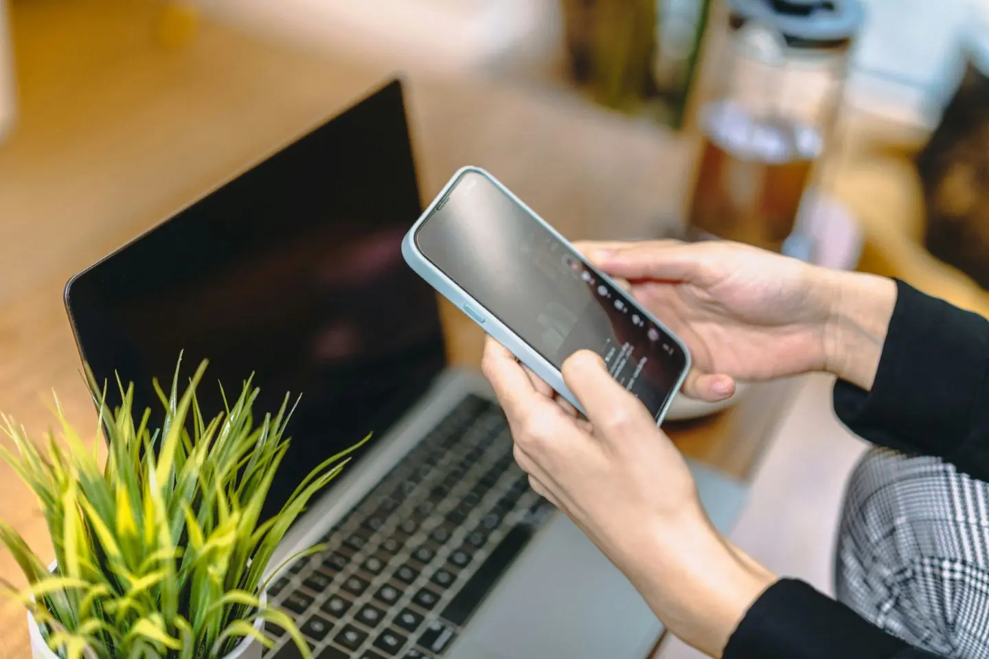 Asmuo naudojasi socialiniu tinklu savo išmaniajame telefone. Nuotraukos fone matomas nešiojamas kompiuteris ir šalia padėtas augalas vazone.