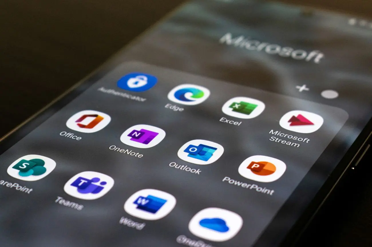 Telefono ekranas su išskleistu programėlių aplanku, pavadinimu "Microsoft". Tarp programėlių yra Authenticator, Edge, Excel, Microsoft Stream, Office, OneNote, Outlook, PowerPoint, Teams, Word ir OneDrive.