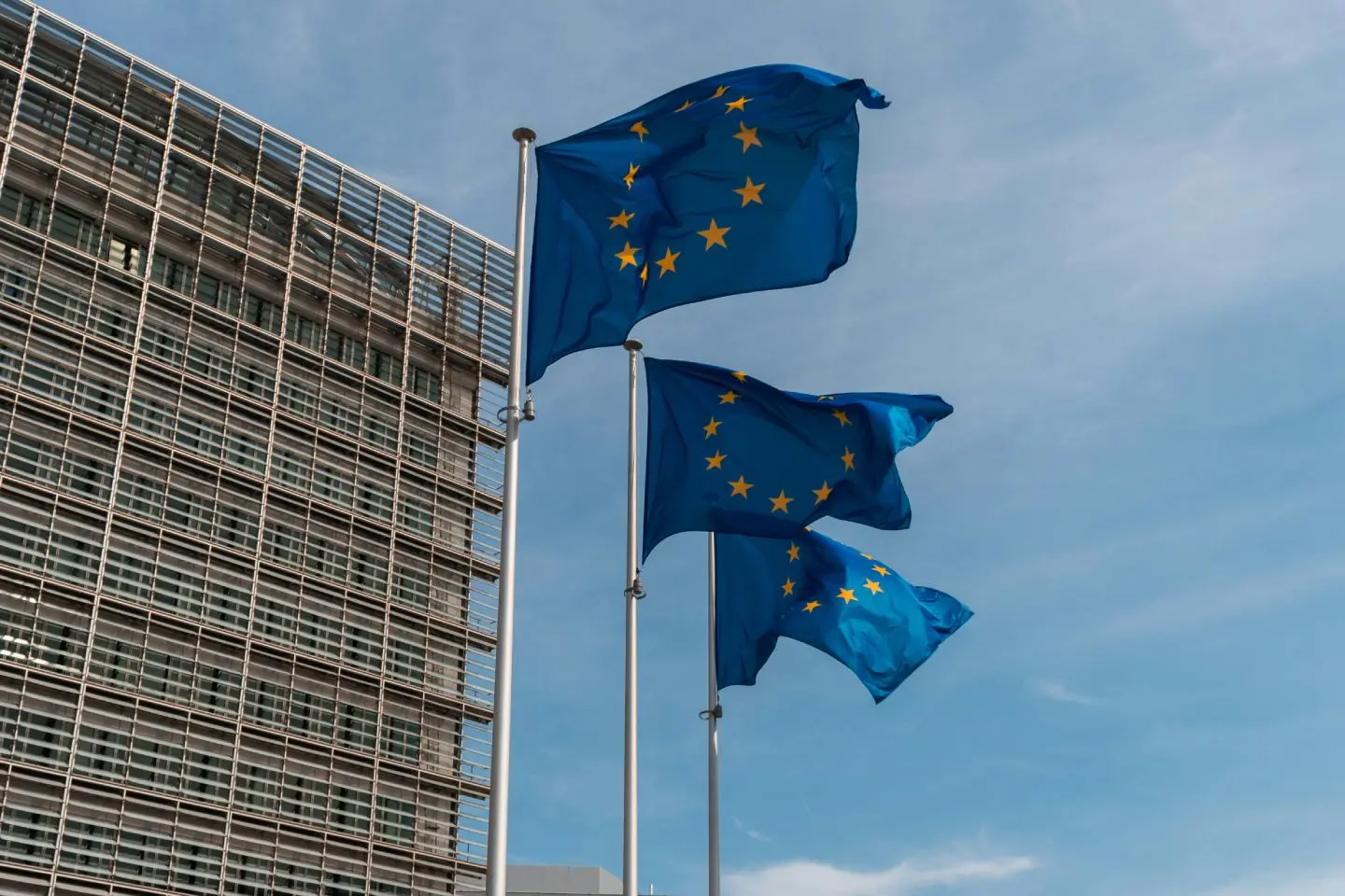 Trys Europos Sąjungos vėliavos plevėsuoja ant stiebų prie modernaus stiklo ir metalo pastato. Fone matomas mėlynas dangus su lengvais debesimis.