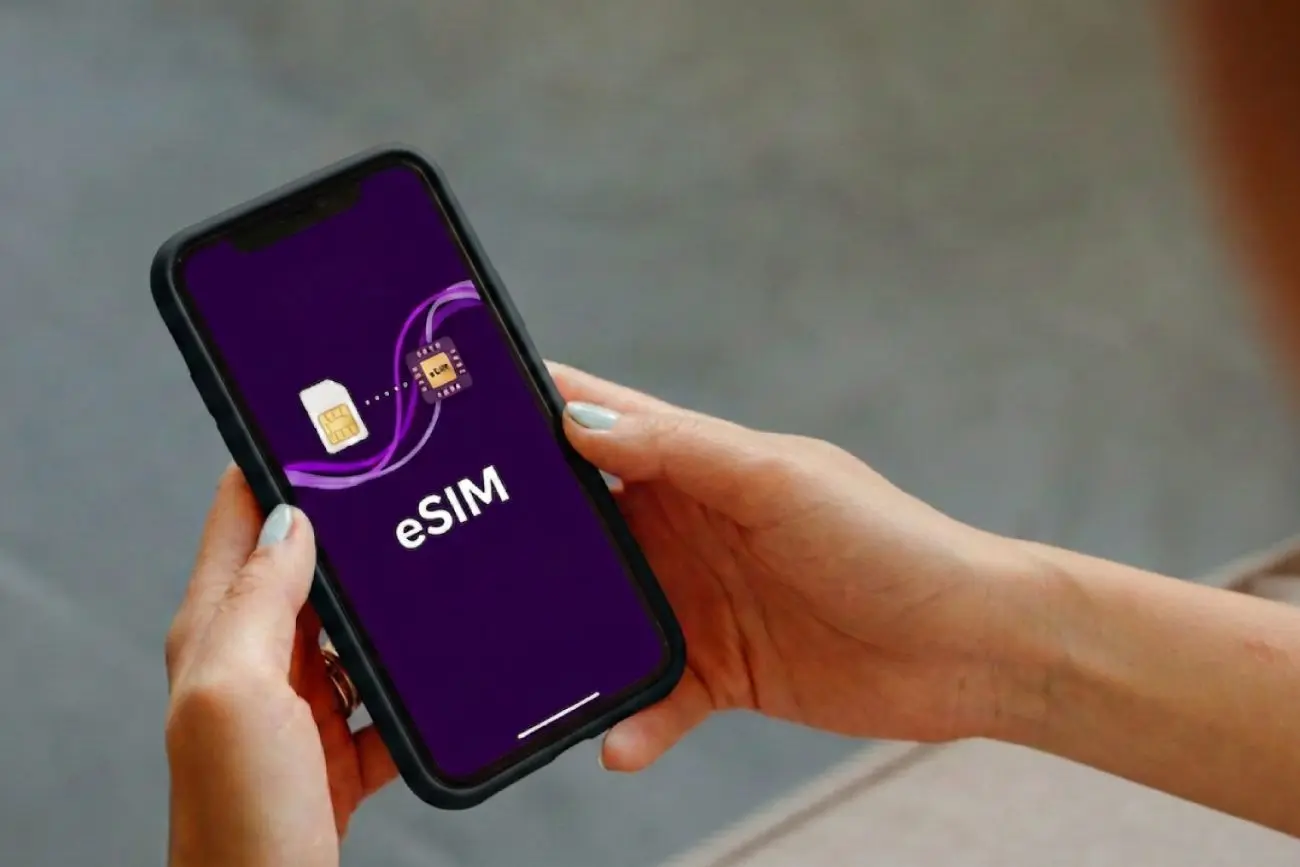 Asmuo laiko išmanųjį telefoną. Ekrane matomas užrašas „eSIM“ ir grafika, vaizduojanti perėjimą nuo fizinės SIM kortelės prie integruoto elektroninio SIM sprendimo.