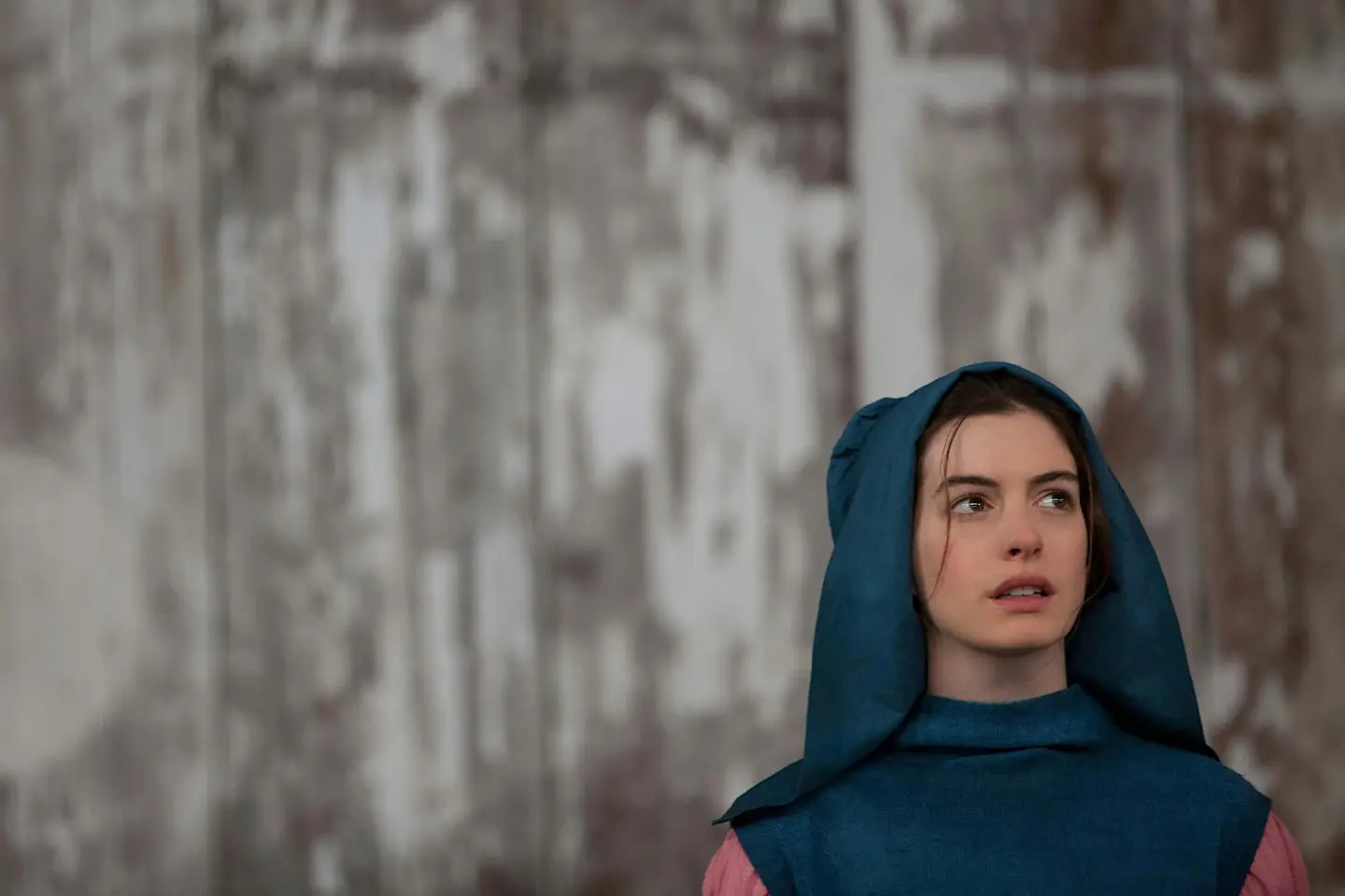 Anne Hathaway kaip Fantina filme „Vargdieniai“ – aktorė su mėlynu gobtuvu žvelgia į tolį priešais nublukusį, tekstūruotą foną