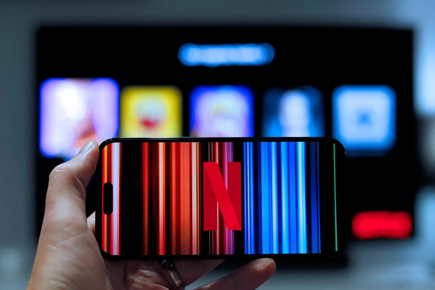 Rankoje laikomas išmanusis telefonas su „Netflix“ logotipu ekrane. Fone matomas neryškus televizoriaus ekranas su srautinio turinio programėlių piktogramomis.