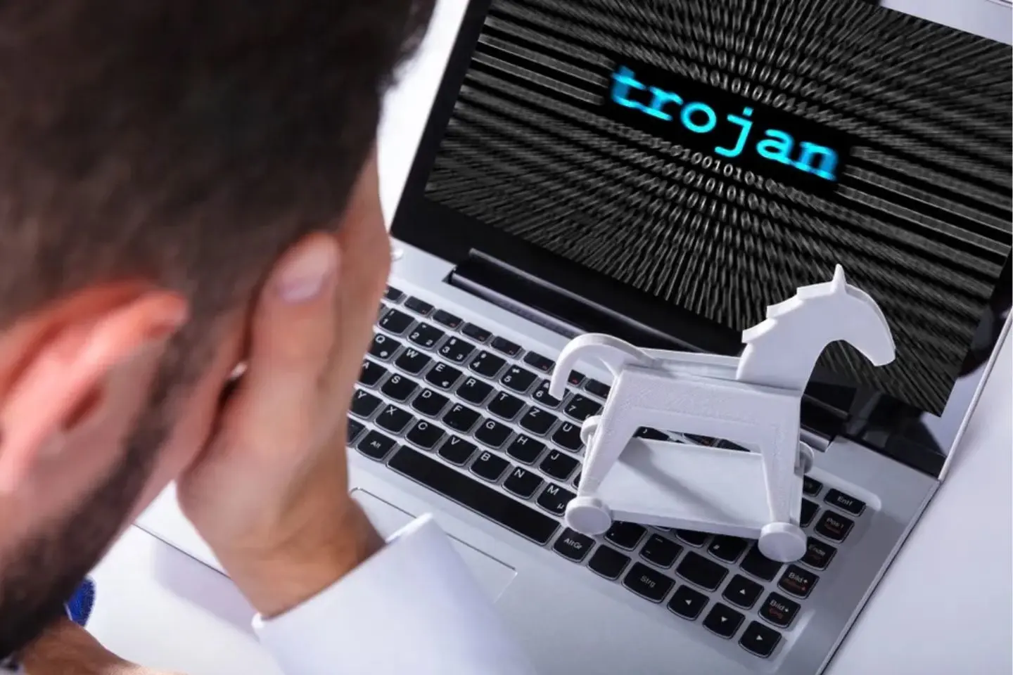 Nešiojamasis kompiuteris su ekrane rodomu užrašu „trojan“. Ant klaviatūros stovi balta Trojos arklio figūrėlė, vaizduojanti kibernetinę grėsmę.