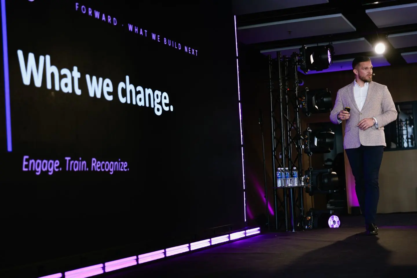 Pranešėjas stovi konferencijos scenoje šalia didelio ekrano. Ekrane matomas tekstas „What we change.“ ir kiti prezentacijos elementai.