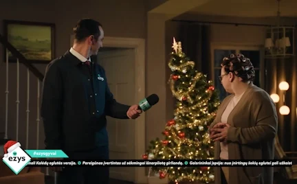Žmogus su mikrofonu kalbina moterį namuose prie judančios šviečiančios Kalėdų eglutės.