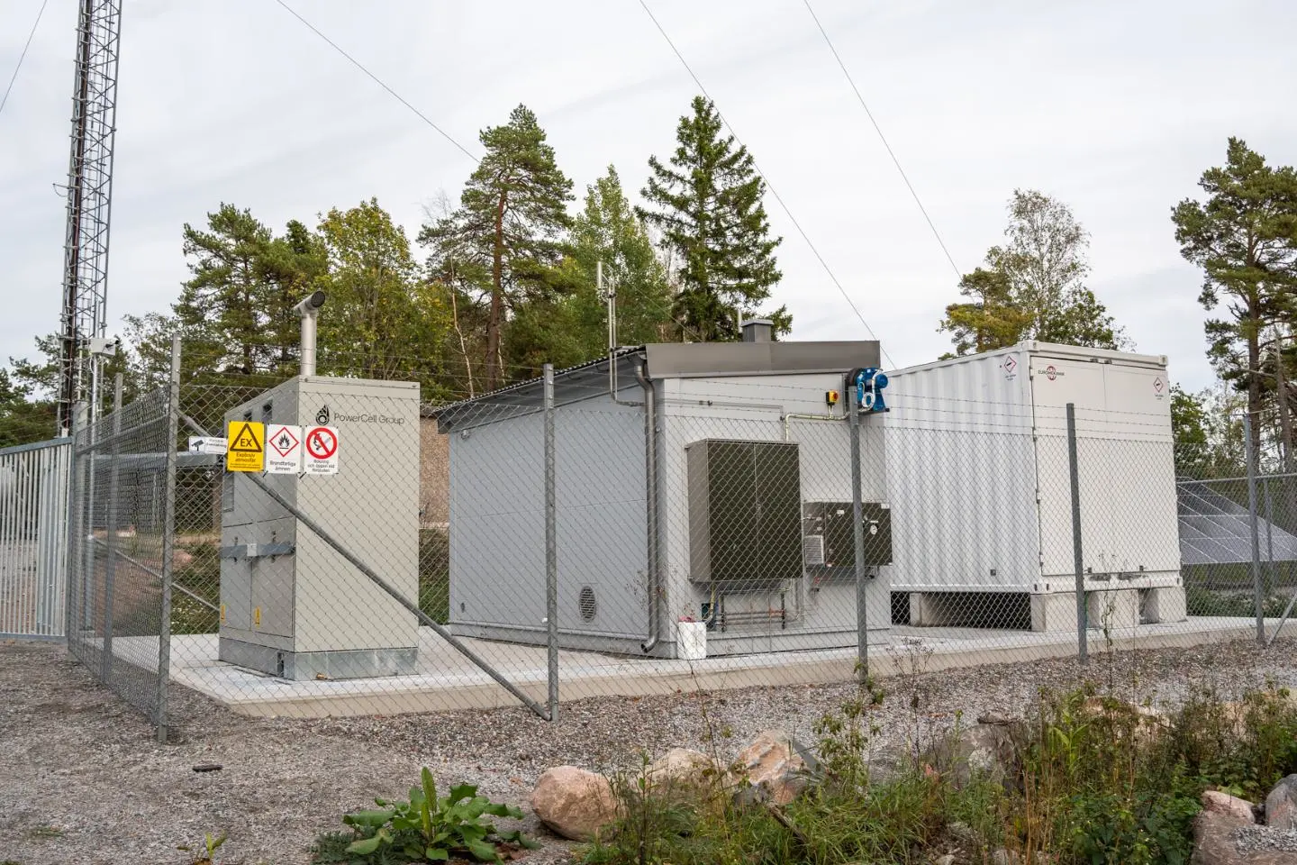 Aptverta techninė vandenilio energijos įrenginių aikštelė miško apsuptyje. Matomi konteineriniai moduliai, elektros įranga, ryšio bokštas ir saugos ženklai ant tvoros.