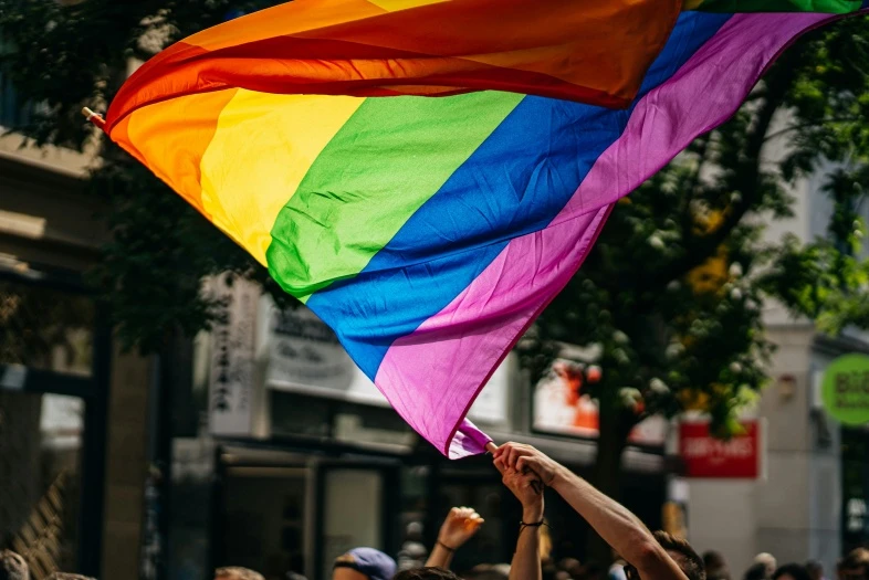 Virš minios lauke plevėsuoja vaivorykštės spalvų Pride vėliava. Fone matomi miesto pastatai ir medžiai.
