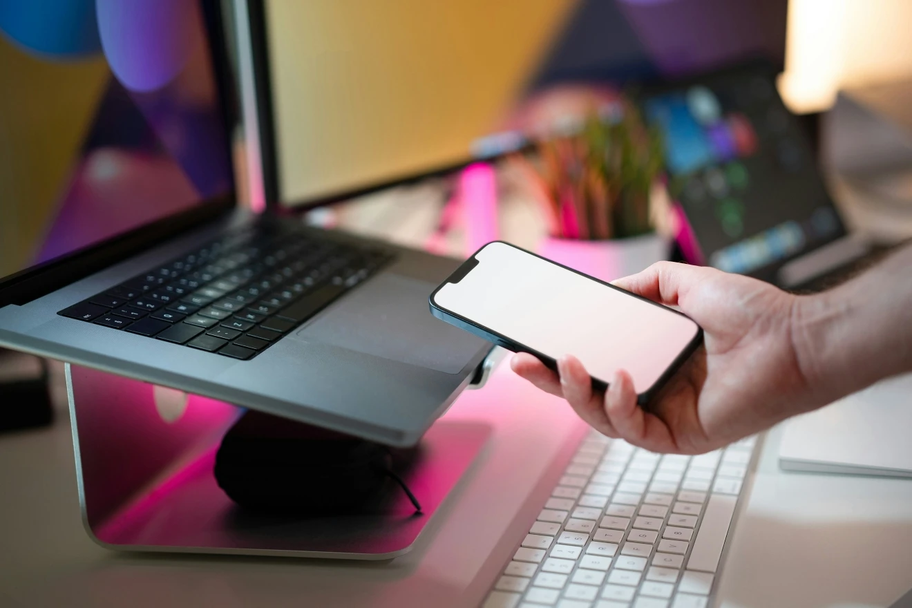 Rankoje laikomas išmanusis telefonas šalia nešiojamojo kompiuterio darbo vietoje. Vaizde pavaizduoti skaitmeniniai įrenginiai ir tarpusavyje susietos technologijos.