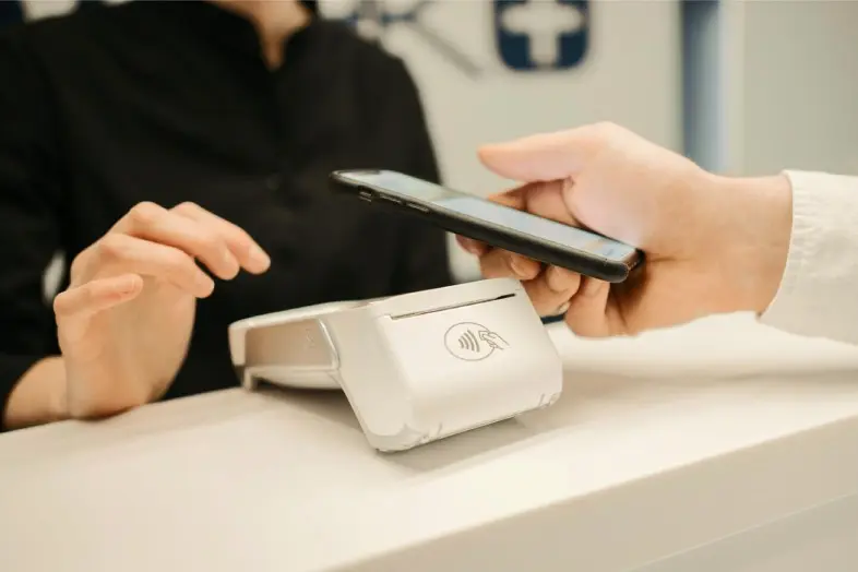 Asmuo laiko išmanųjį telefoną virš bekontakčio mokėjimo terminalo. Vaizduojamas atsiskaitymas telefonu naudojant NFC technologiją.