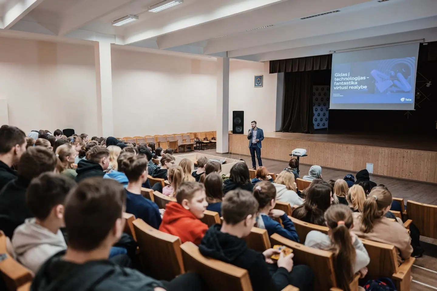 Auditorijoje susirinkę moksleiviai klausosi pranešėjo scenoje. Ekrane rodomas pristatymas apie technologijas ir virtualią realybę.