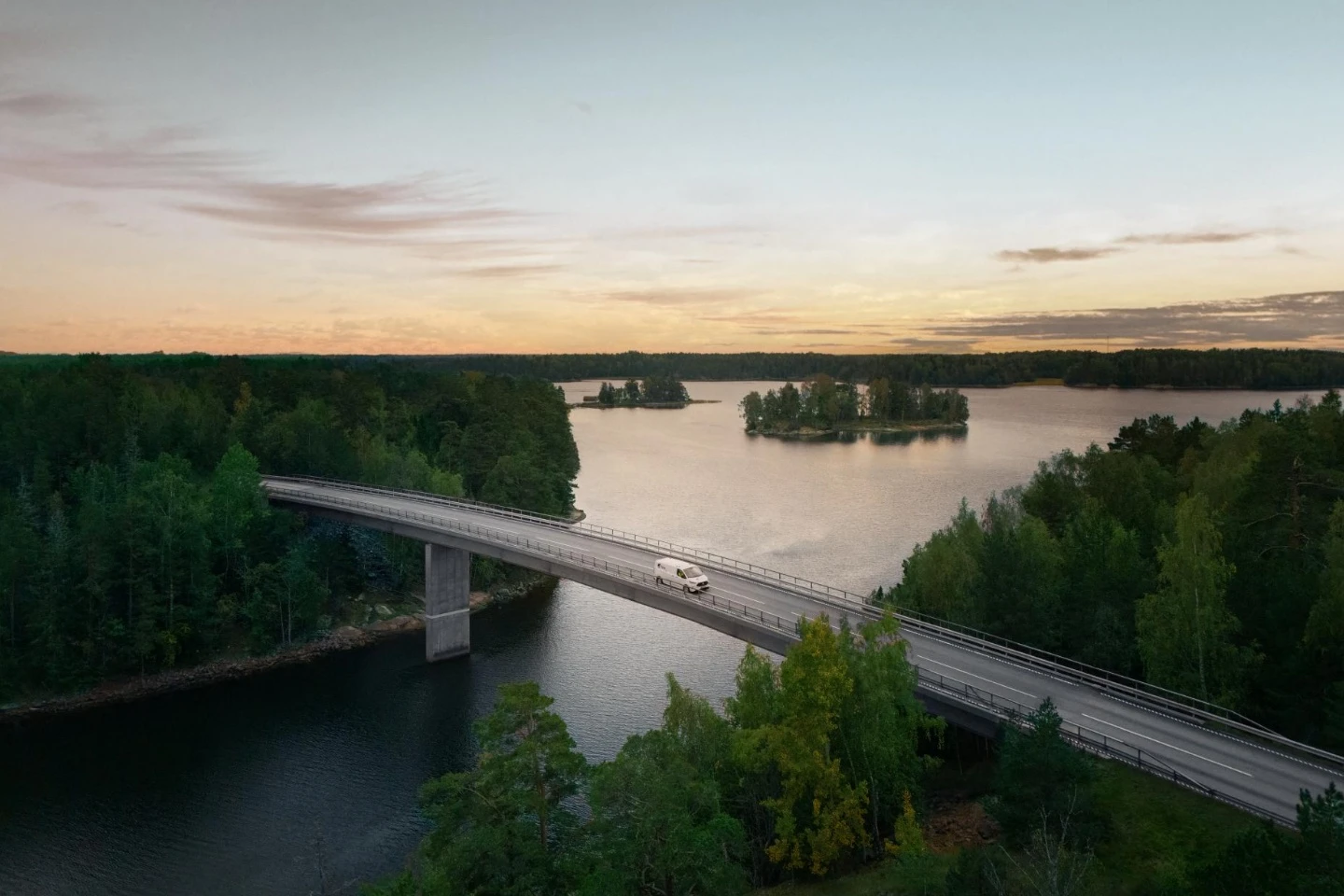 Vaizdas iš oro: tiltu per ežerą važiuoja baltas Telia automobilis. Aplink matyti miškingos salos, vandens telkiniai ir vakaro dangus.