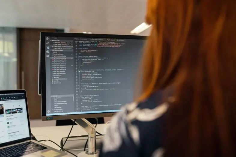 Asmuo sėdi prie darbo vietos ir žiūri į didelį monitorių su atidarytu programavimo kodu. Ant stalo matomas nešiojamasis kompiuteris.