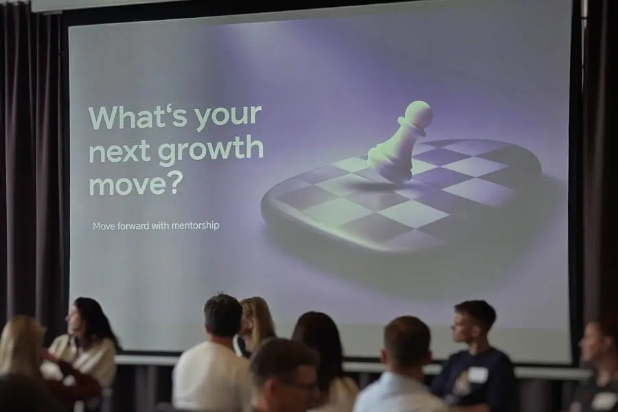Auditorijoje susirinkę žmonės klausosi pranešimo. Dideliame ekrane rodoma mentorystės tema „What’s your next growth move?“ ir šachmatų lentos vaizdas.