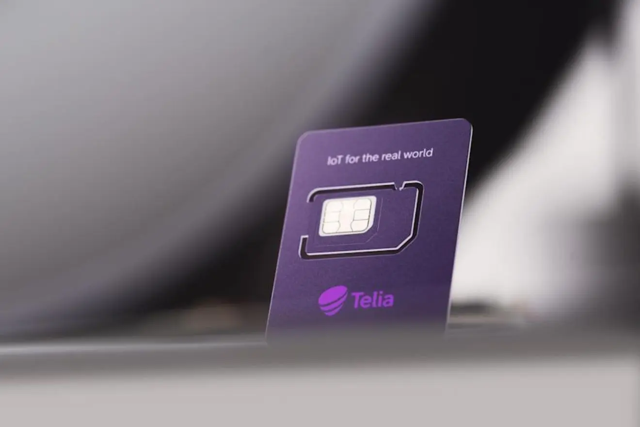 Violetinė „Telia“ IoT SIM kortelė stovi vertikaliai išblukusiame fone. Ant kortelės matomas SIM lustas ir užrašas „IoT for the real world“.