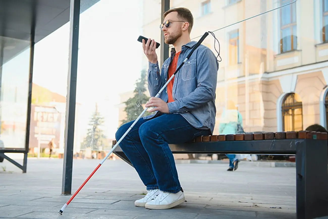 Vyras su tamsiais akiniais sėdi lauke ant suoliuko ir kalba į telefoną. Prie suoliuko atremta balta orientacinė lazdelė, aplink matoma miesto aplinka.