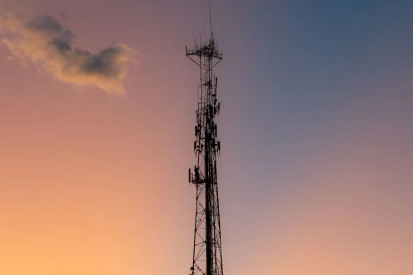 Aukštas telekomunikacijų bokštas su antenomis stovi prieš spalvotą vakaro dangų. Horizontas nuspalvintas šiltais saulėlydžio atspalviais.