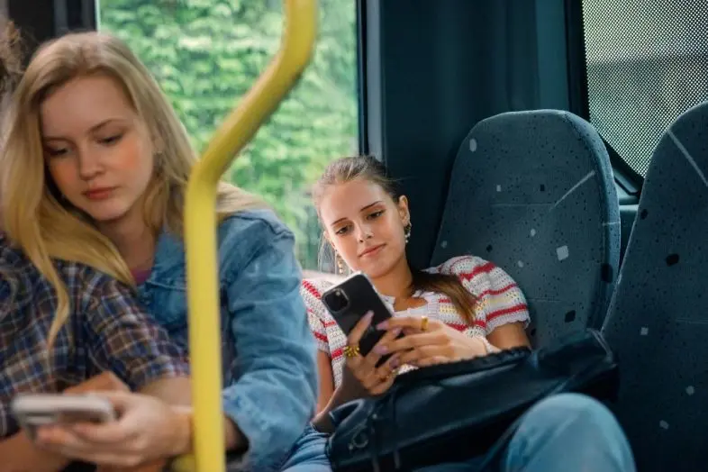 Autobuso salone dvi merginos naudojasi mobiliaisiais telefonais. Viena mergina sėdi priekyje, kita ilsisi sėdynėje prie lango ir žiūri į telefoną.
