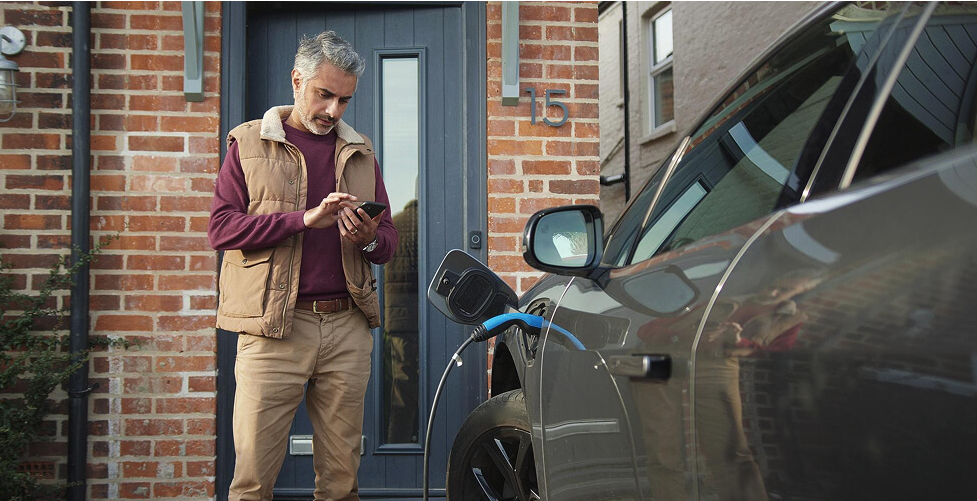 Žilas vyras stovi prie kraunamo elekromobilio ir žiūri į telefoną