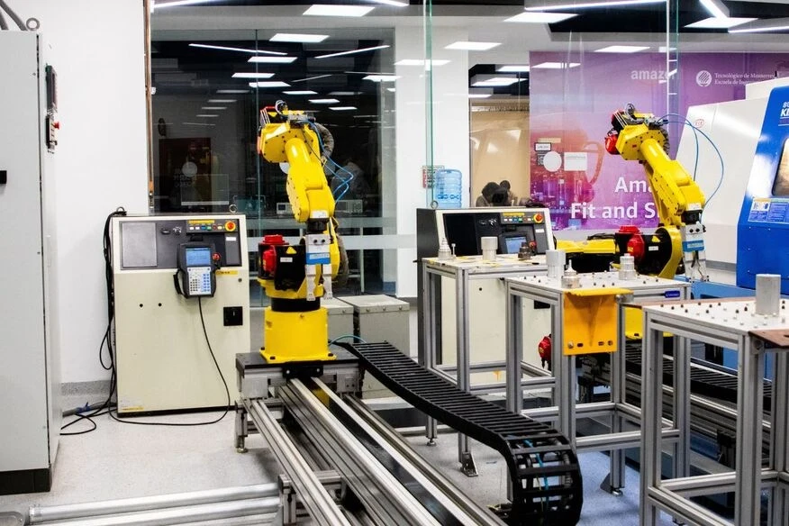 Automatizuotos gamybos laboratorija su dviem pramoniniais robotais. Robotų rankos dirba prie surinkimo stotelių, apsuptų valdymo ir transportavimo įrangos.