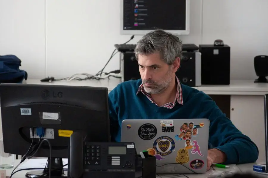Biuro aplinkoje prie darbo vietos sėdintis asmuo naudojasi nešiojamuoju kompiuteriu. Ant stalo matomi monitorius, telefonas ir kitos darbo priemonės.