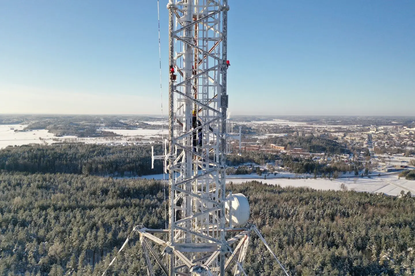 Mobiliojo ryšio bokštas žiemiškame kraštovaizdyje. Bokšto konstrukcija padengta šerkšnu, o aplink matyti miškas, laukai ir miesto teritorija.