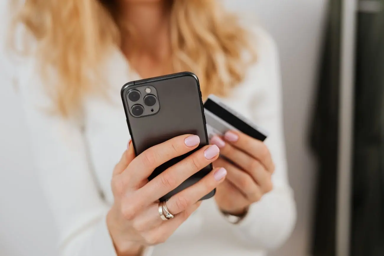 Moteris laiko išmanųjį telefoną vienoje rankoje, o banko kortelę kitoje. Nuotrauka vaizduoja mobilųjį atsiskaitymą ar apsipirkimą internete.