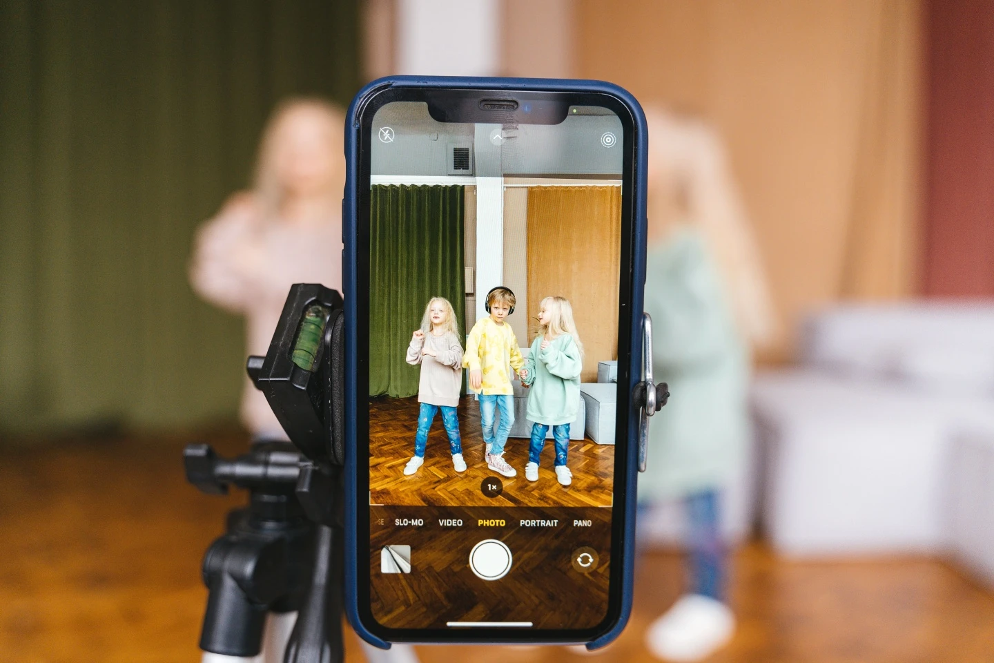 Išmanusis telefonas ant stovo filmuoja tris vaikus patalpoje. Telefono ekrane matomas fotografavimo režimas ir filmuojami vaikai, o fonas už telefono yra išlietas.